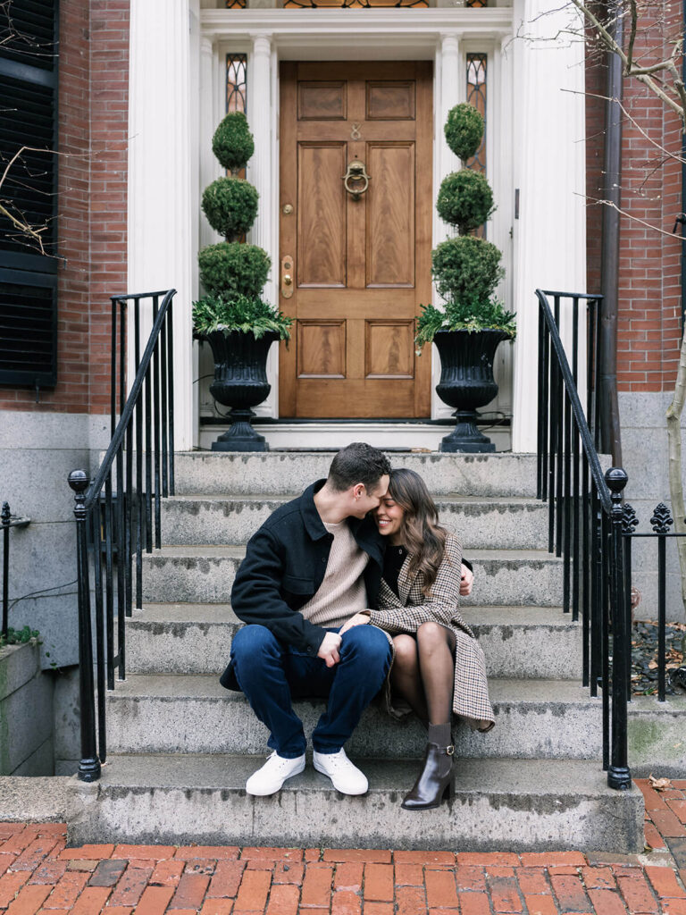 Couple sits on the steps of a brick townhouse on a Beacon Hill street during engagement photos