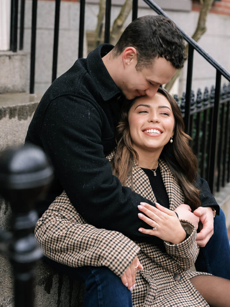 Groom kisses bride's temple while sitting on stone steps on a Beacon Hill street during engagement photos