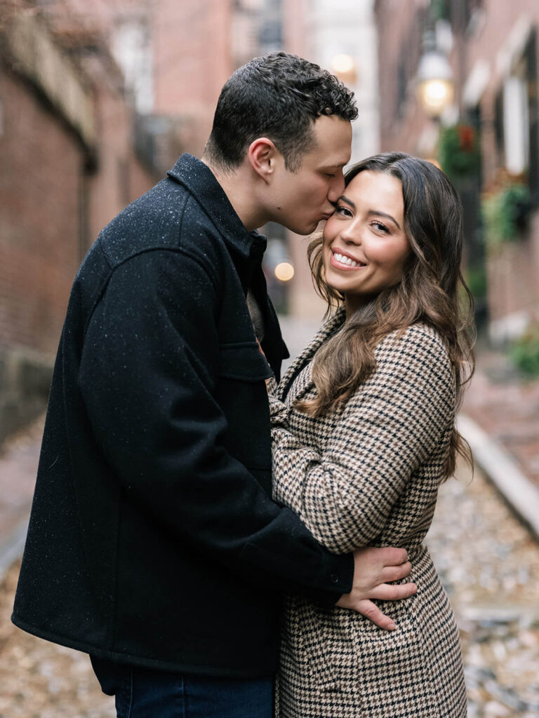 He kisses her forehead while wrapping both arms around her on Acorn Street cobblestones in Beacon Hill