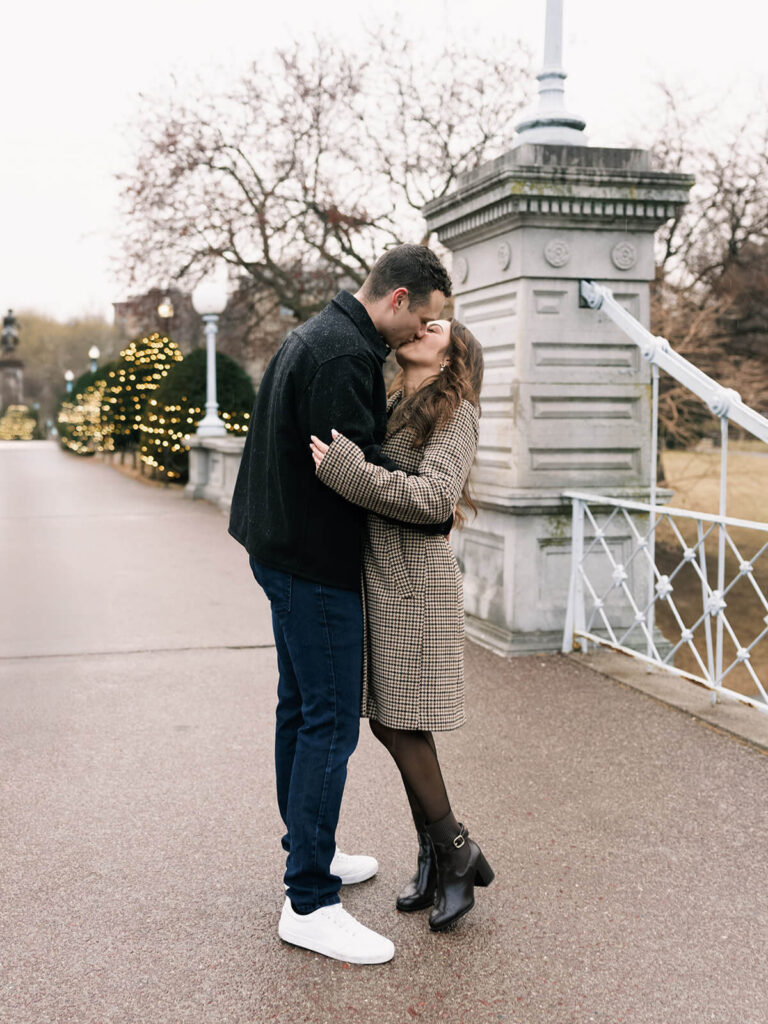 Couple kisses while standing near a stone pillar at a pedestrian bridge in Boston Public Garden