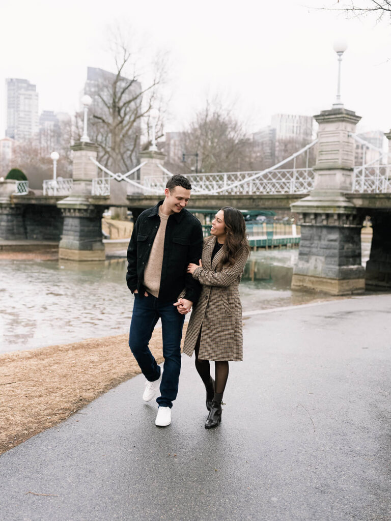 Alyssa and Jason walk hand-in-hand along a path beside a lagoon with bridge in background in Boston Public Garden