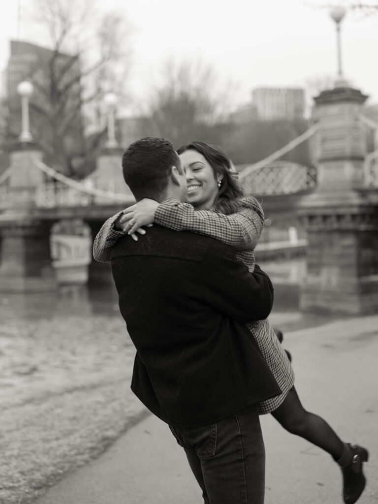 Groom lifts bride off the ground in an embrace beside the lagoon in Boston Public Garden