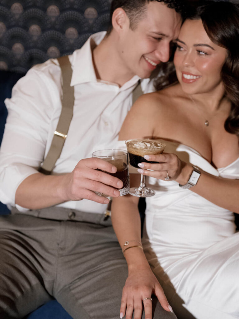 Couple clinking cocktail glasses on velvet booth at 89 Charles during Boston engagement session