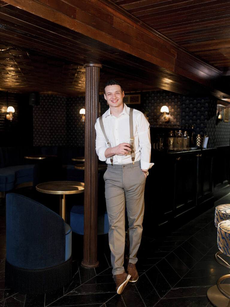 Man in white shirt and suspenders leaning against pillar at cocktail lounge engagement session