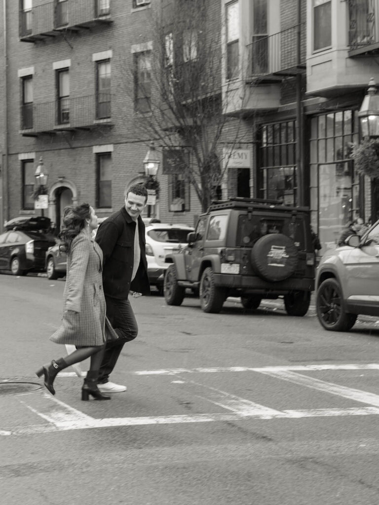 Couple crossing city street together in black and white with brick buildings behind them