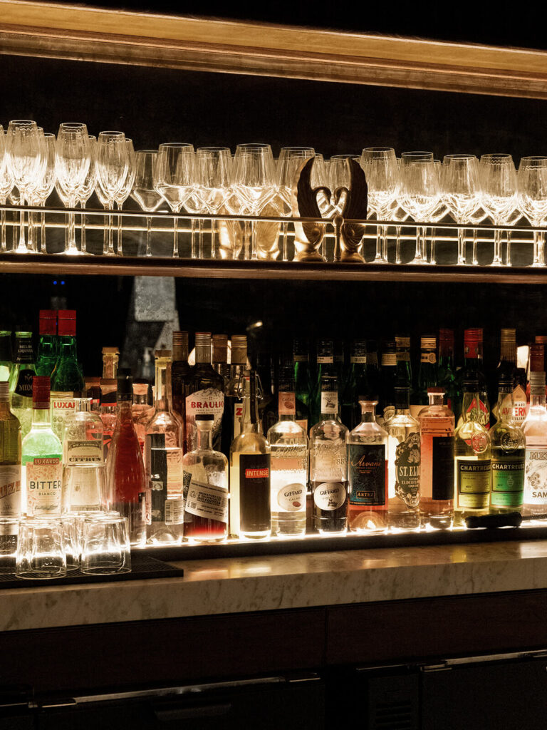 Backlit liquor bottles and wine glasses on shelves behind cocktail bar detail shot