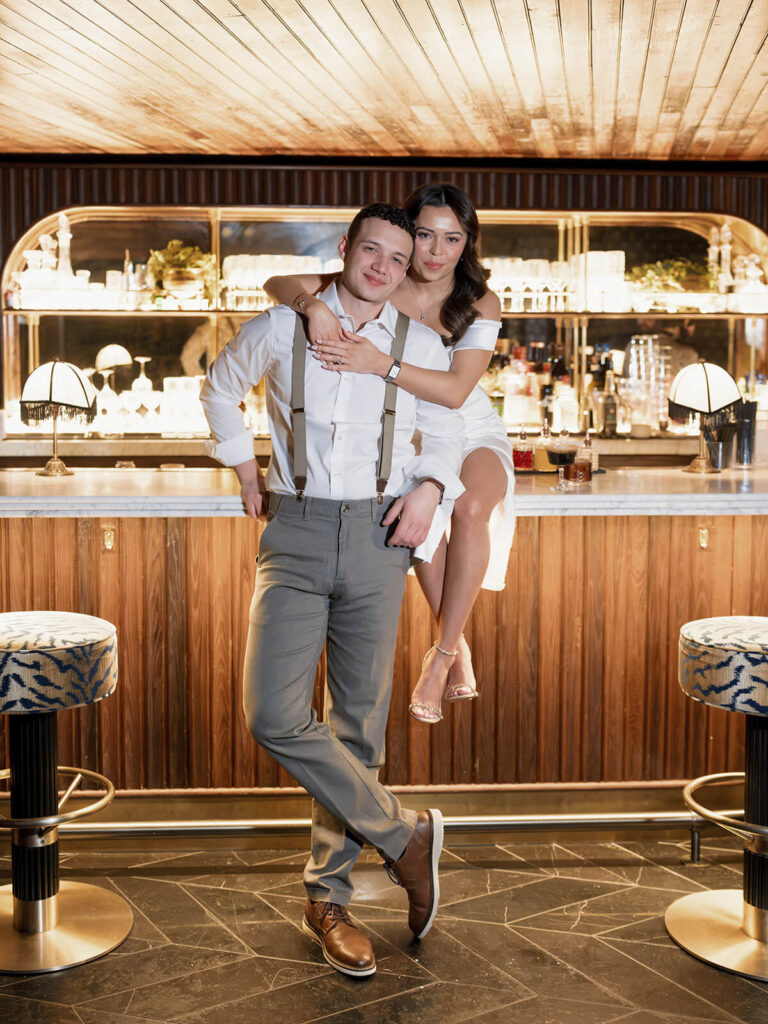 Man carrying woman piggyback-style in front of bar both smiling during engagement photos
