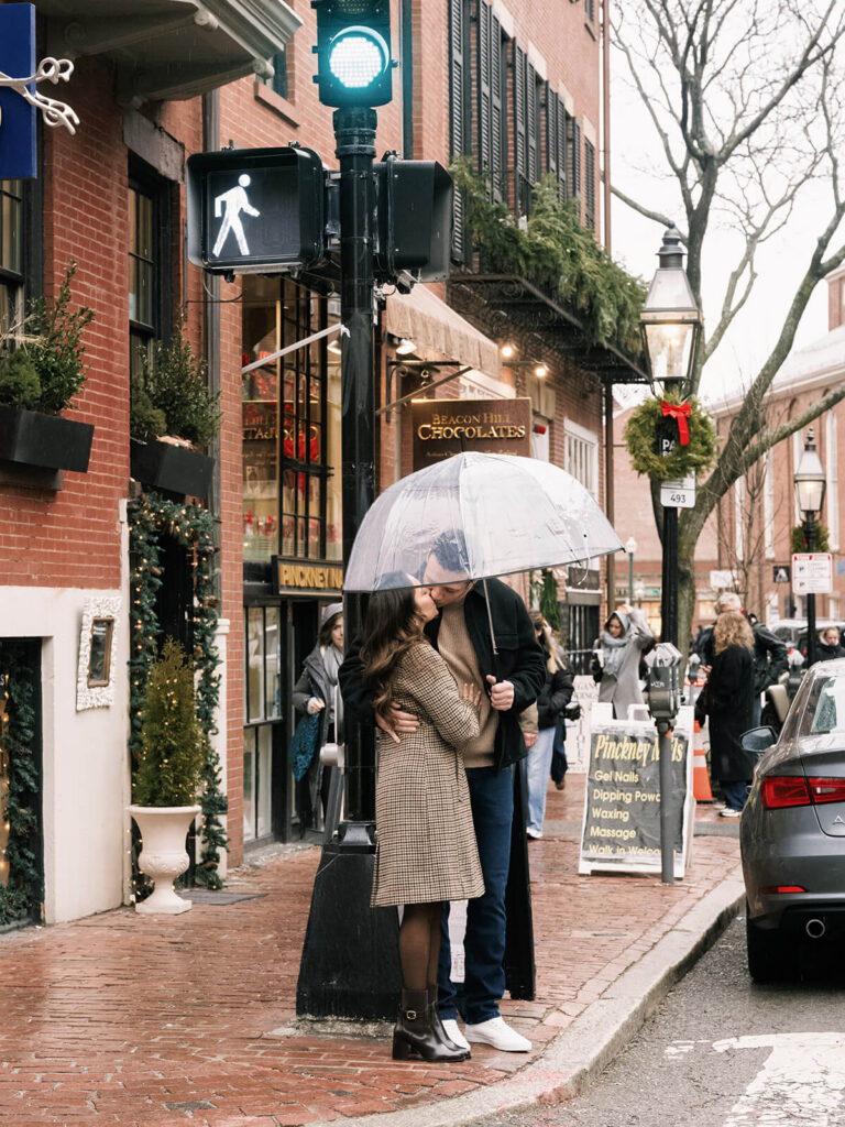 Couple kissing under clear umbrella on rainy Beacon Hill street with holiday decorations