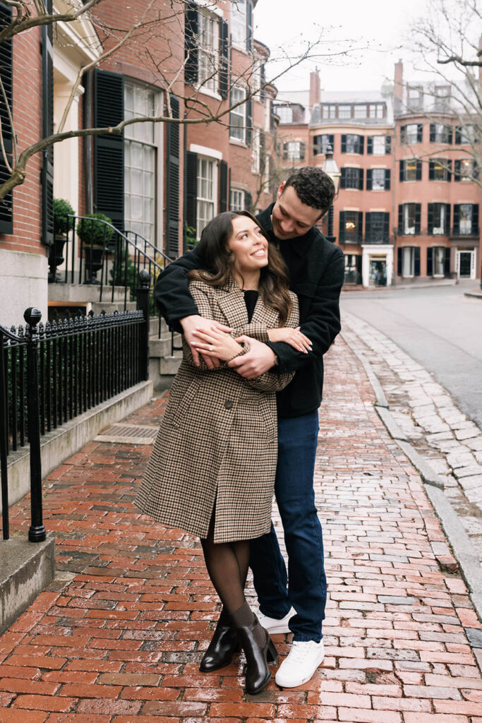 Couple embracing and smiling at camera on cobblestone street with brick townhouses in black and white