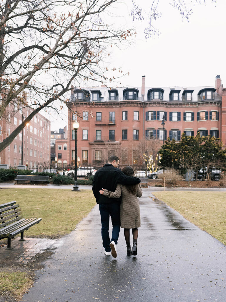 Couple walking away with arms around each other on wet park path with brick buildings behind