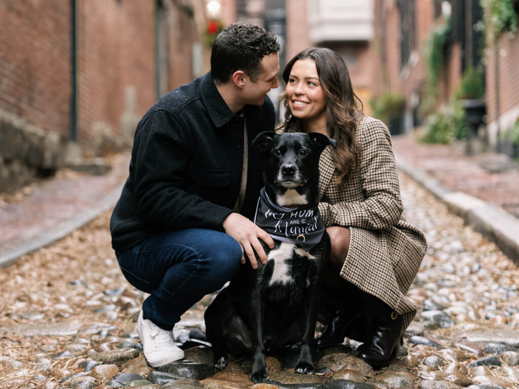 Couple crouching with dog wearing My Humans Are Getting Married bandana on cobblestone alley