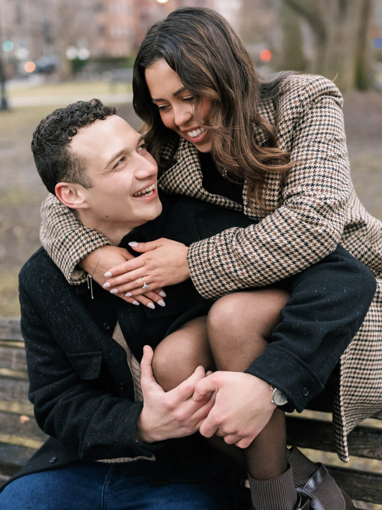 Close-up of couple laughing on park bench woman embracing man with engagement ring visible