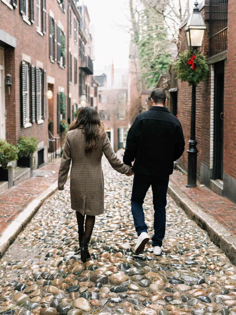 Couple walking hand-in-hand down cobblestone alley with brick buildings and holiday wreaths
