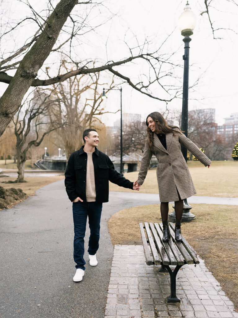 Woman walking on park bench holding hands with man on pathway both smiling with city behind