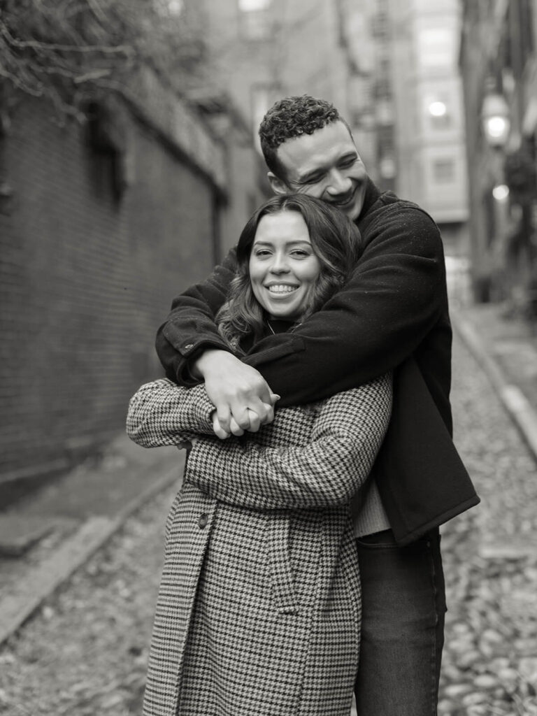 Man hugging woman from behind on cobblestone alley with brick buildings black and white