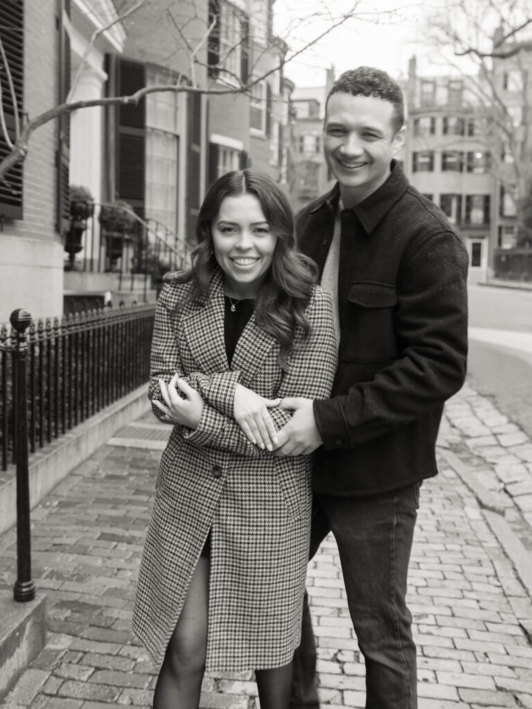 Couple embracing and smiling on cobblestone street with brick townhouses black and white