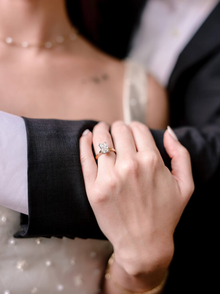 Close-up of emerald-cut diamond engagement ring on gold band resting on suited arm Charlotte session
