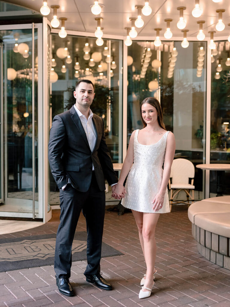 Couple standing hand-in-hand facing camera outside restaurant with globe light canopy Charlotte