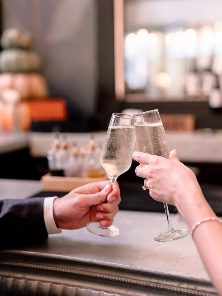 Close-up of two hands toasting champagne flutes at bar showing engagement ring Charlotte