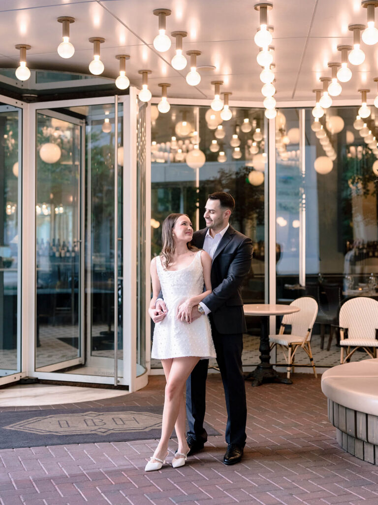 Man embracing woman from behind under globe lights at restaurant entrance Charlotte engagement session