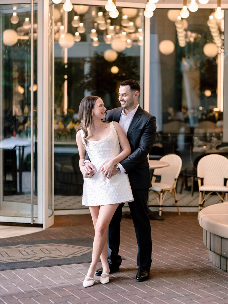 Couple laughing in embrace outside restaurant with globe lights uptown Charlotte engagement photos