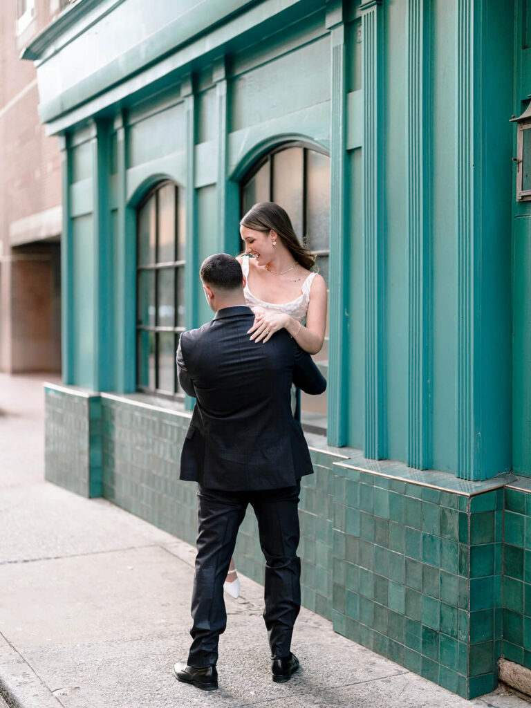 Man lifting laughing woman against teal building exterior on Charlotte city sidewalk
