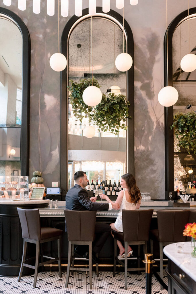 Couple seated at bar toasting champagne with arched mirrors and globe lights overhead Charlotte