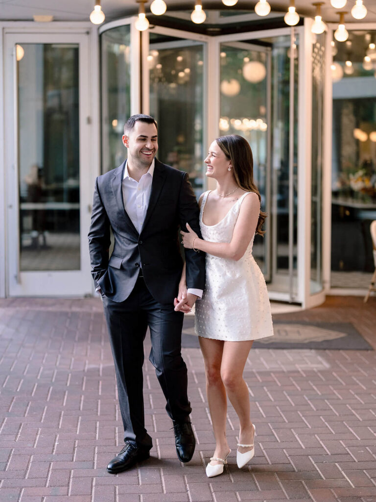 Couple walking arm-in-arm laughing on brick patio near restaurant Charlotte engagement session