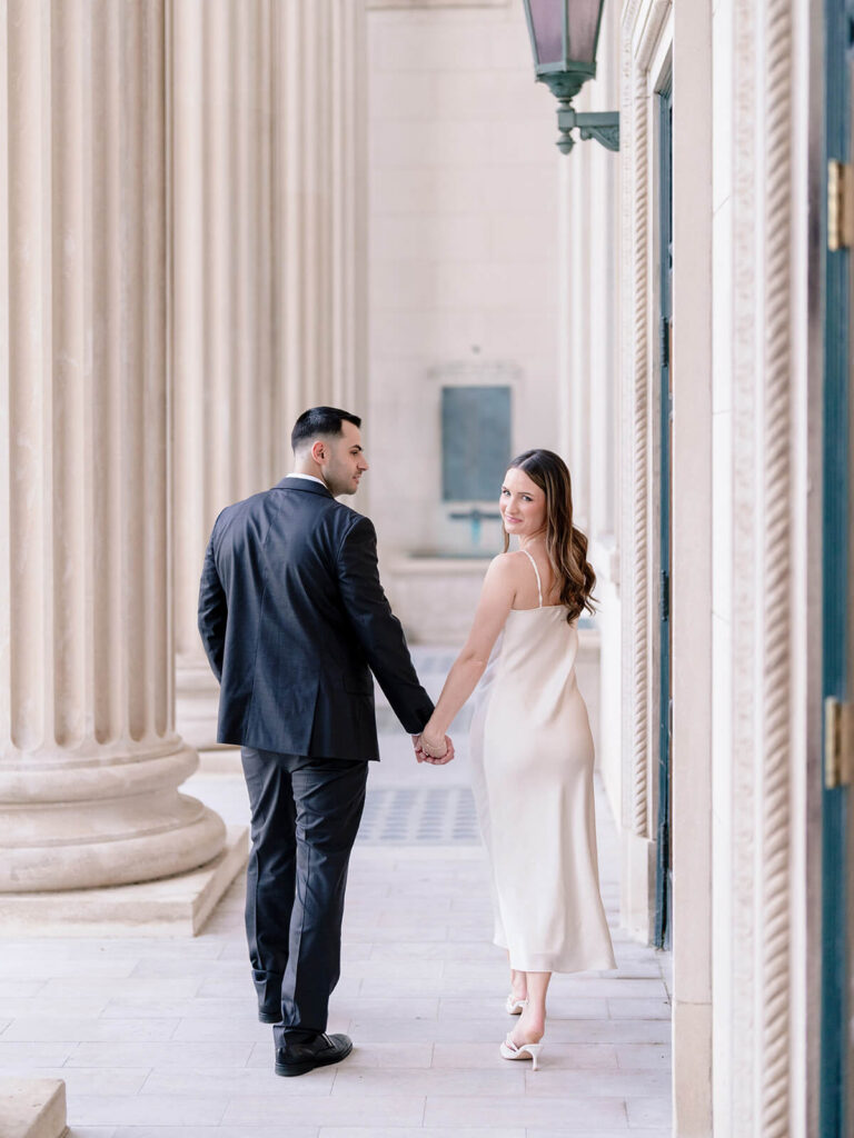 Couple holding hands walking through classical colonnade woman looks back at camera Gandhi Park