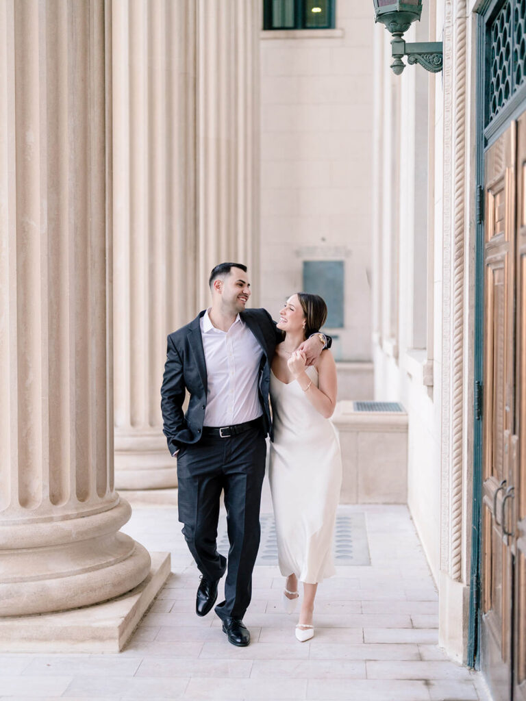 Couple walking arm-in-arm through columned portico laughing Gandhi Park Charlotte engagement photos