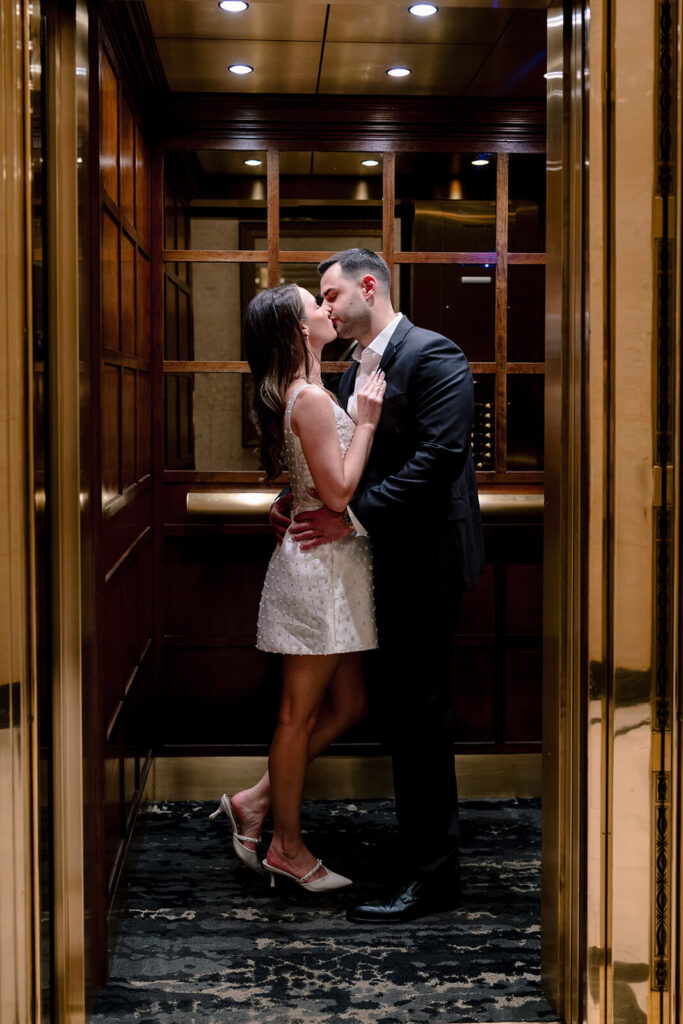 Couple kissing inside wood-paneled elevator with brass doors and mirrored walls at hotel