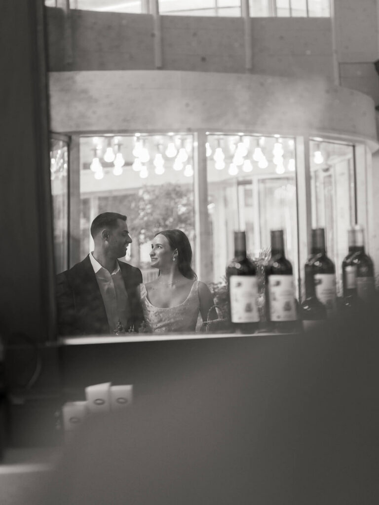 Black and white of couple looking at each other through bar mirror wine bottles in foreground
