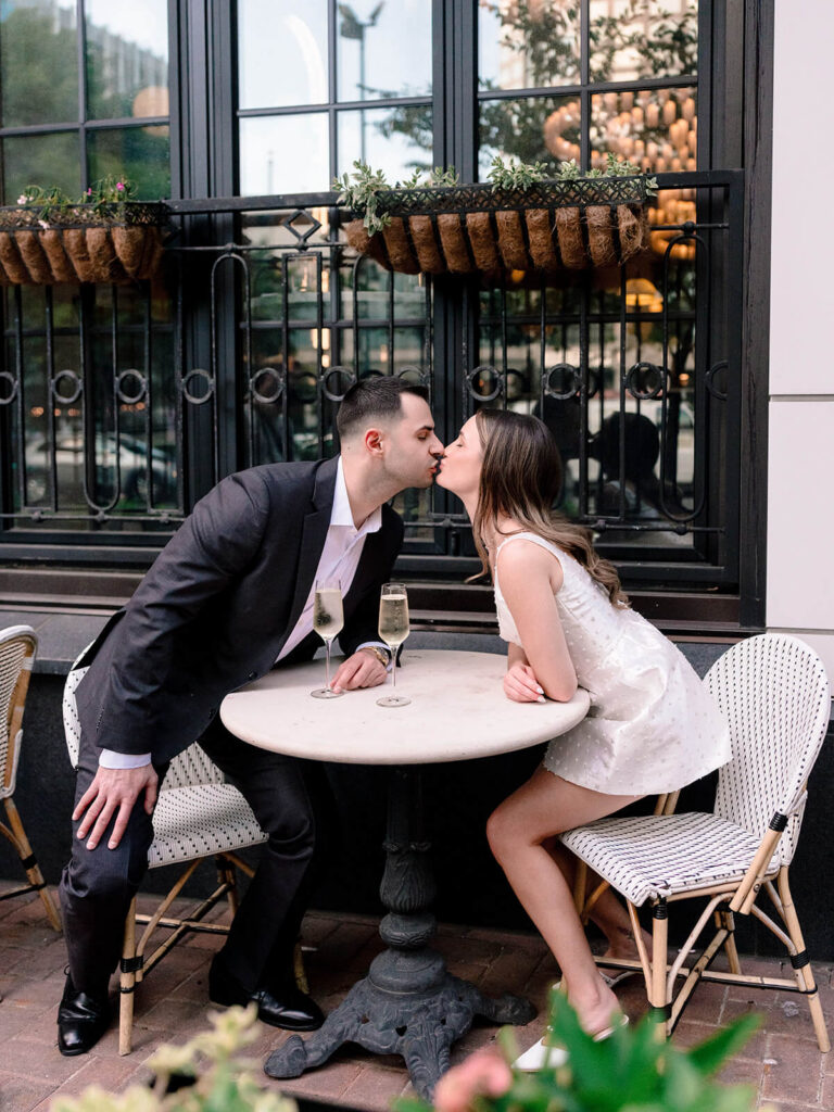 Couple kissing across bistro table with champagne glasses at outdoor cafe patio Charlotte
