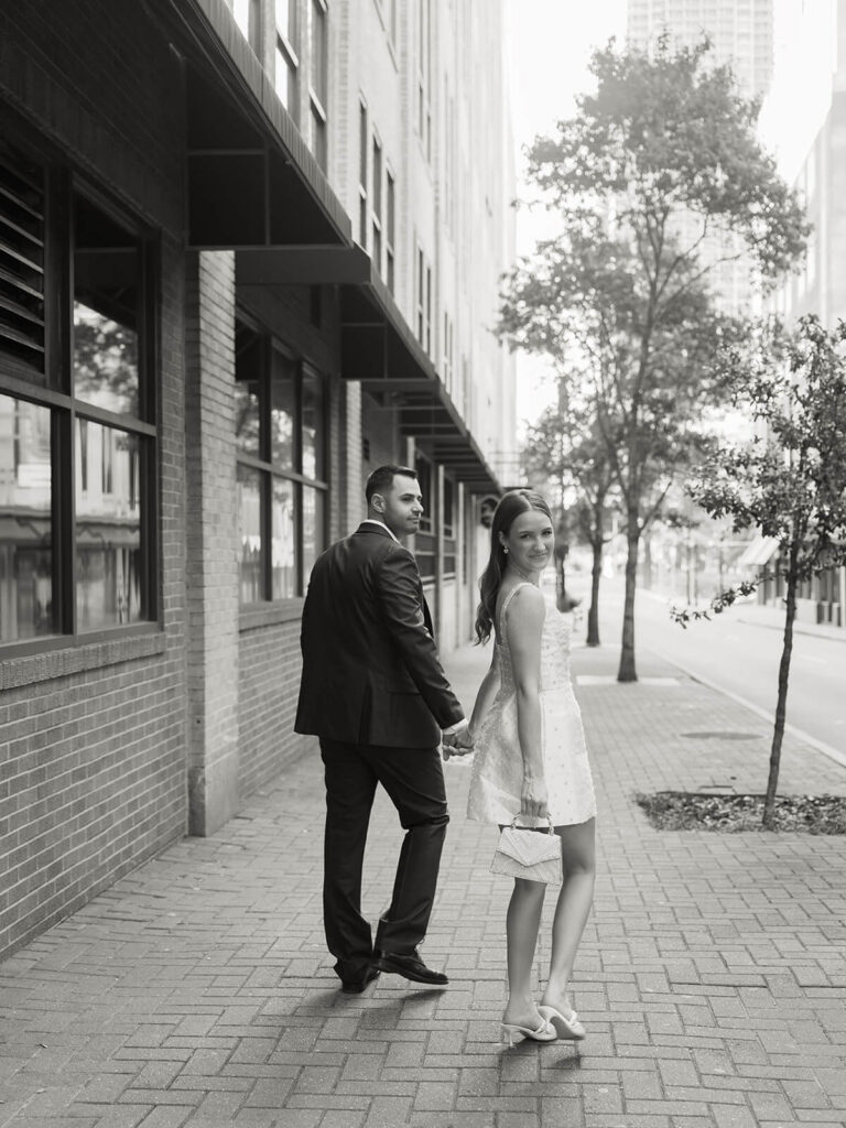 Couple looking back over shoulders holding hands on brick sidewalk downtown Charlotte engagement