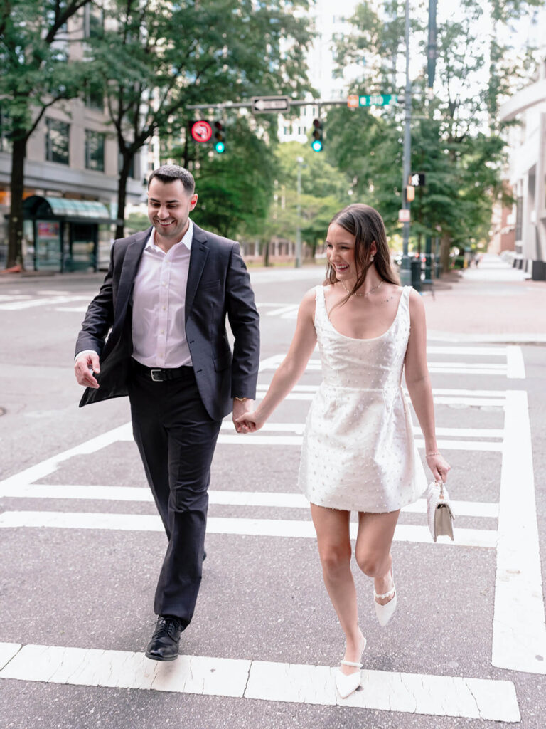 Couple holding hands crossing downtown Charlotte crosswalk laughing with trees in background