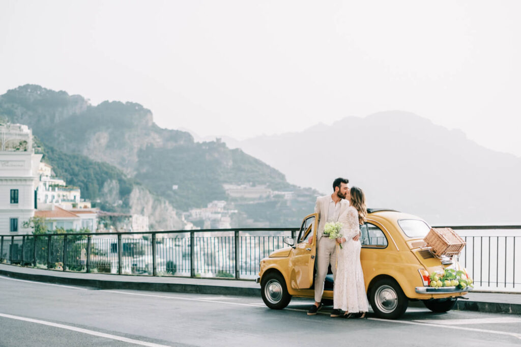 Alessandra and Alessio sharing a sweet kiss next to their Fiat, framed by Amalfi’s iconic cliffs and seaside charm.