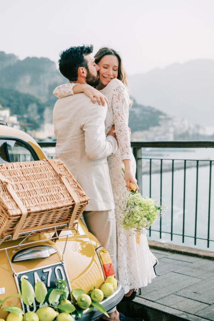 Alessandra smiling as Alessio embraces her, with the Amalfi Coast and their lemon-adorned Fiat in the background.