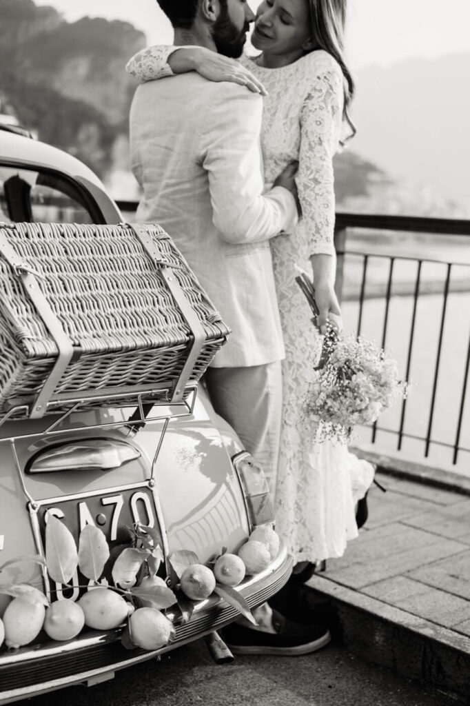 Black-and-white detail of Alessandra leaning into Alessio, clutching a bouquet next to their vintage yellow Fiat.