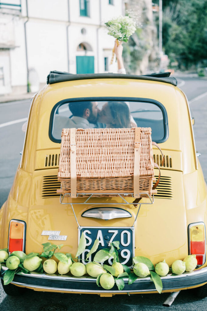 Rear window view of Alessandra and Alessio sharing a kiss inside their Fiat, wicker picnic basket fastened to the trunk.