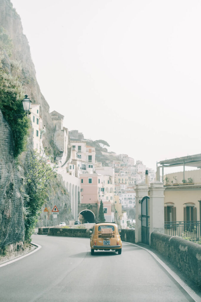 Vintage yellow Fiat cruising along a winding coastal road in Amalfi, with colorful cliffside architecture in the distance.