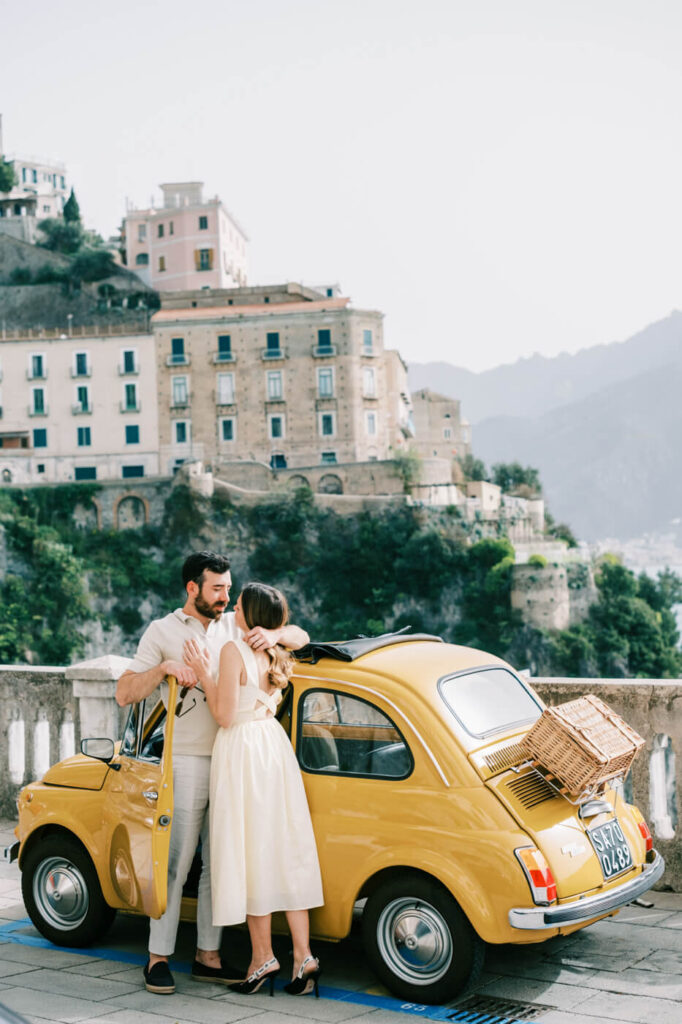 Alessandra and Alessio sharing a tender moment beside their vintage Fiat, framed by the grand architecture of Amalfi.