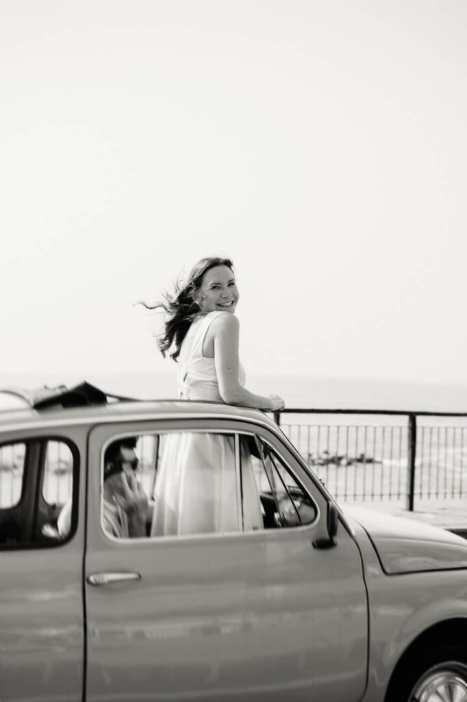 Black-and-white photo of Alessandra smiling from the sunroof of a yellow Fiat, capturing the carefree spirit of the Amalfi Coast.