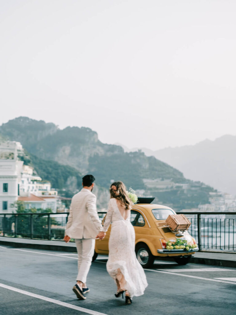 Alessandra and Alessio walking hand in hand next to their yellow Fiat, set against the cliffs of the Amalfi Coast.