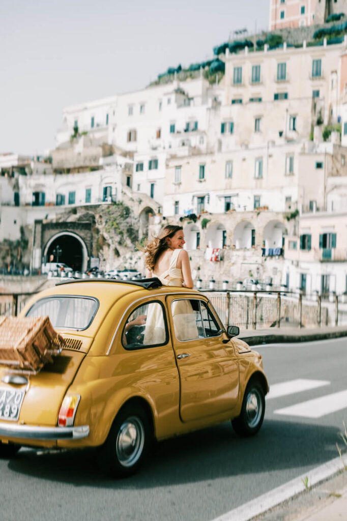 Alessandra leaning out of the vintage Fiat while driving through Amalfi’s scenic streets, with pastel buildings rising behind her.