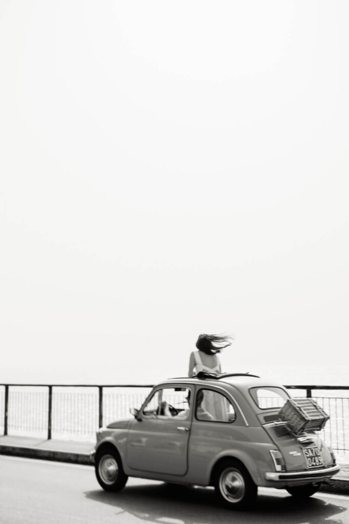 Black-and-white photo of Alessandra standing through the Fiat’s sunroof, her hair blowing in the breeze along the Amalfi Coast.