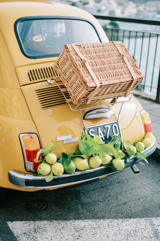 Close-up of a yellow vintage Fiat’s trunk, featuring a wicker picnic basket and Amalfi lemons for a charming coastal detail.