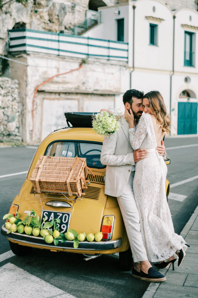 Alessandra and Alessio leaning against their lemon-adorned Fiat, sharing an intimate moment along the Amalfi Coast.