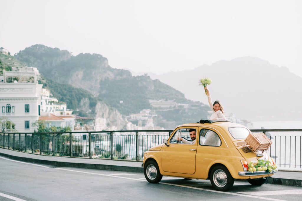 Alessandra raising her bouquet in celebration through the sunroof of a yellow Fiat, overlooking the panoramic Amalfi coastline.