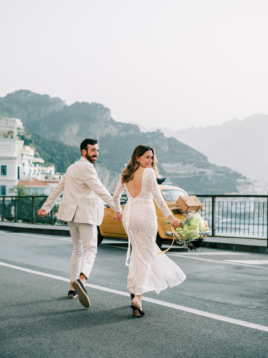 Featured image of couples session with Alessandra smiling over her shoulder while holding Alessio’s hand, running towards their vintage Fiat on the Amalfi Coast.