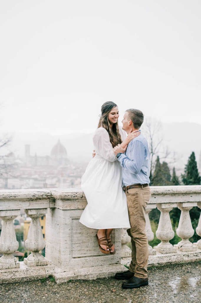 Candid engagement moment in Florence, Italy – The bride sits playfully on the edge of a stone balcony while the groom gazes up at her, the historic city visible in the distance.